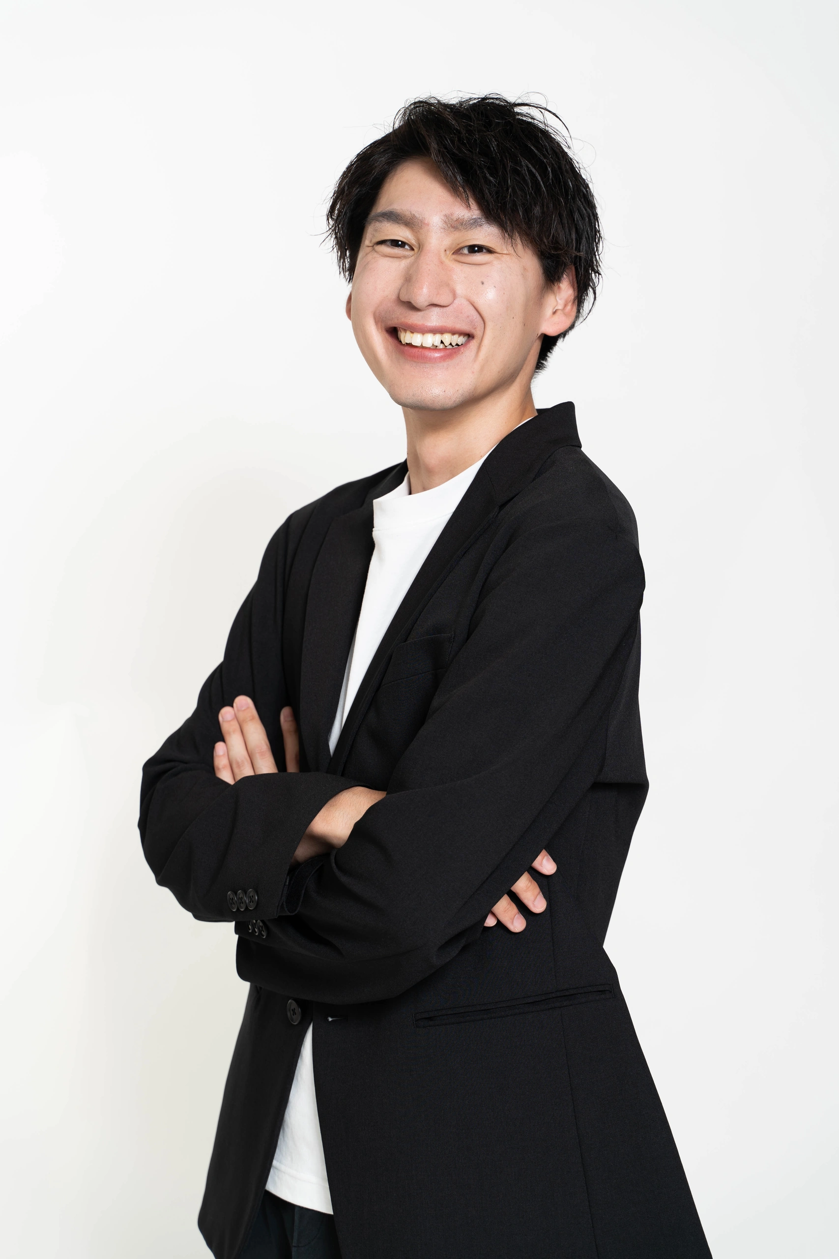 白い背景で腕を組み笑顔を見せる黒いジャケットの人物